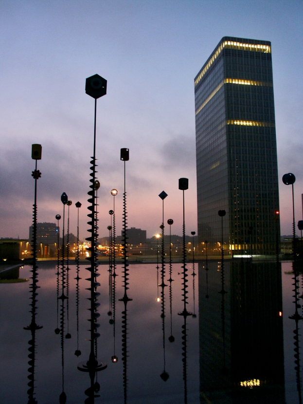 Mehrere Masten und Gebäude mit Lichtern unter einem bewölkten Himmel, mit Spiegelungen der Masten