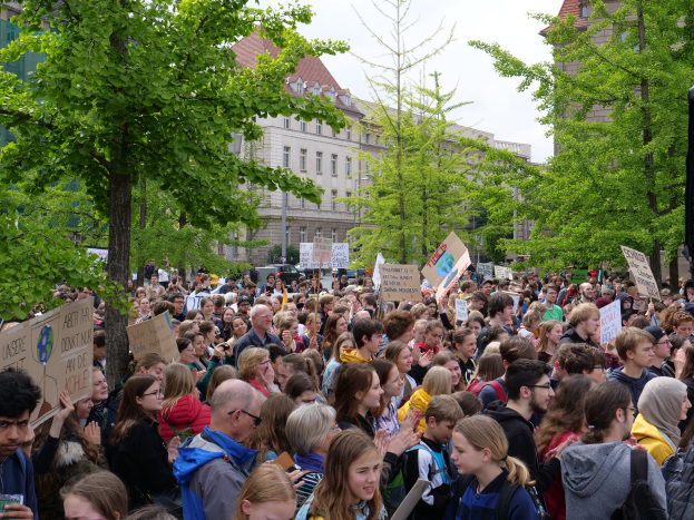 Eine große Menschenmenge mit Schildern steht vor einem Gebäude in Berlin, mit Bäumen, Fahrzeugen und einem Lautsprecher zu sehen, unter einem klaren Himmel.