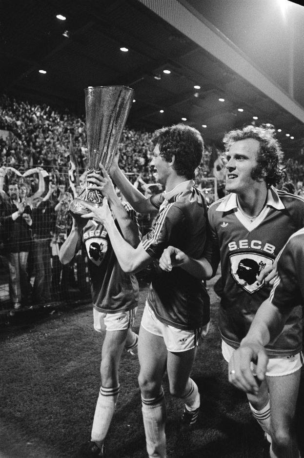 Schwarzes und weißes Bild von Männern, die mit einem Pokal auf einem Fußballfeld feiern, umgeben von einer Menge hinter einem Zaun unter Stadionbeleuchtung.