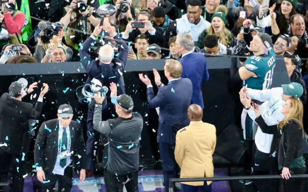 Philadelphia Eagles Spieler feiern mit dem Vince Lombardi Trophy nach dem Gewinn des Super Bowls, einige halten den Pokal und andere werfen Konfetti, mit einem Geländer und Fotografen im Hintergrund.