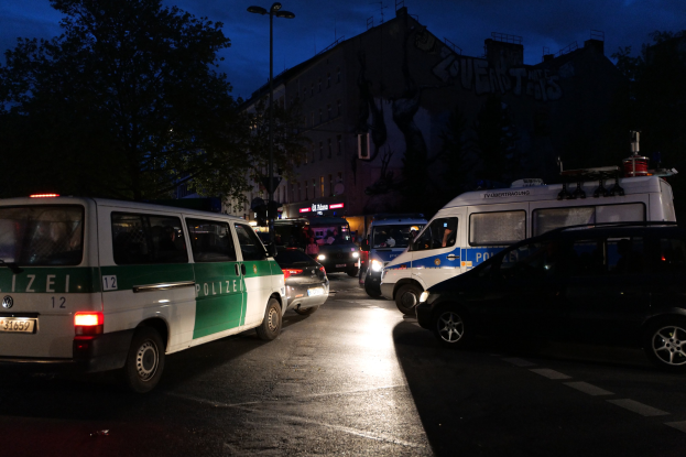 Eine Gruppe von Polizeiwagen, darunter ein Van, ein Auto und ein Rad, parkt nachts am Straßenrand, mit Gebäuden, Bäumen, Laternen und einem Sternenhimmel im Hintergrund.