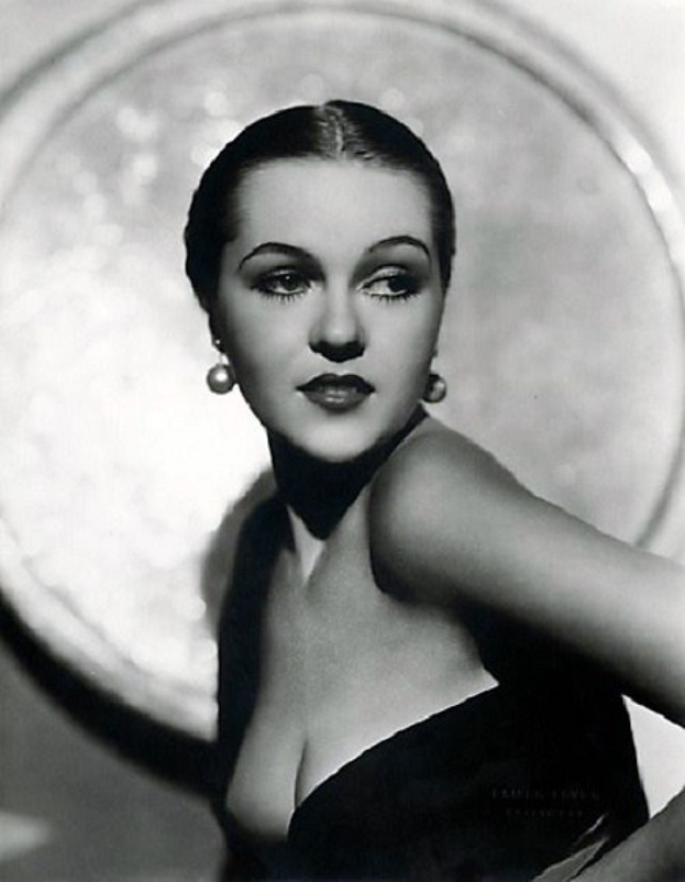 Schwarz-weißes Porträt einer Frau in einem langärmligen, hochgeschlossenen Kleid mit 1940er-Jahre-Stil und Make-up, die selbstbewusst in die Kamera schaut mit einem unscharfen Hintergrund.