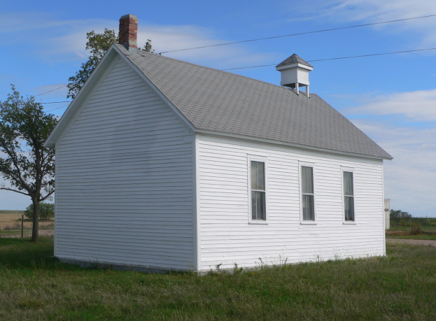 Eine kleine weiße Schule in einer grünen Wiese mit ein paar Bäumen und einem klaren blauen Himmel.