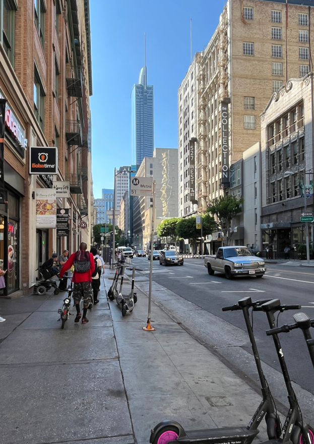 Eine Gruppe von Menschen, die auf Roller durch eine Stadtstraße fahren, gesäumt von hohen Gebäuden, mit einem geparkten Pick-up, Straßenlaternen, sichtbarem Himmel, Bäumen und Schildern.