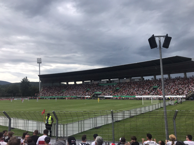 Große Zuschauermenge bei einem Fußballspiel in einem bewölkten, offenen Stadion mit beleuchteten Masten, umgeben von einem Zaun, Bäumen und Hügeln im Hintergrund.