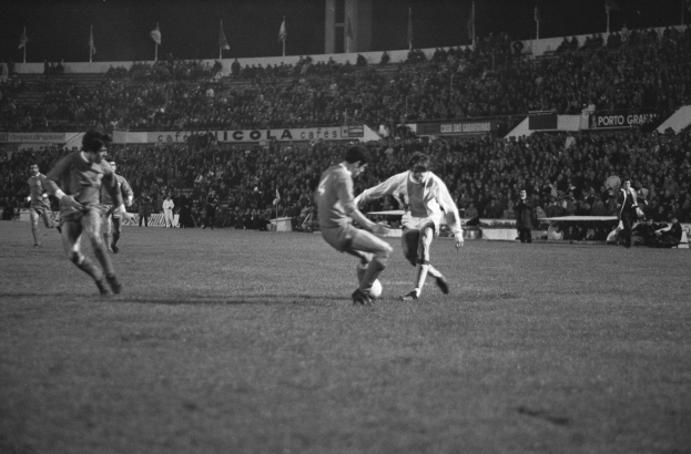 Schwarzes und weißes Bild eines Fußballspiels mit Spielern auf einem Feld und Zuschauern auf den Tribünen, mit Fahnen im Hintergrund.