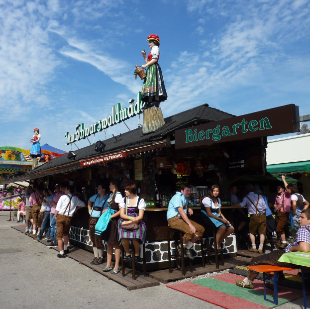 Group of people in front of a building at Oktoberfest in Munich, some in traditional Bavarian clothing, others seated on stools with tables of bottles and a board in the background under trees and cloudy sky.