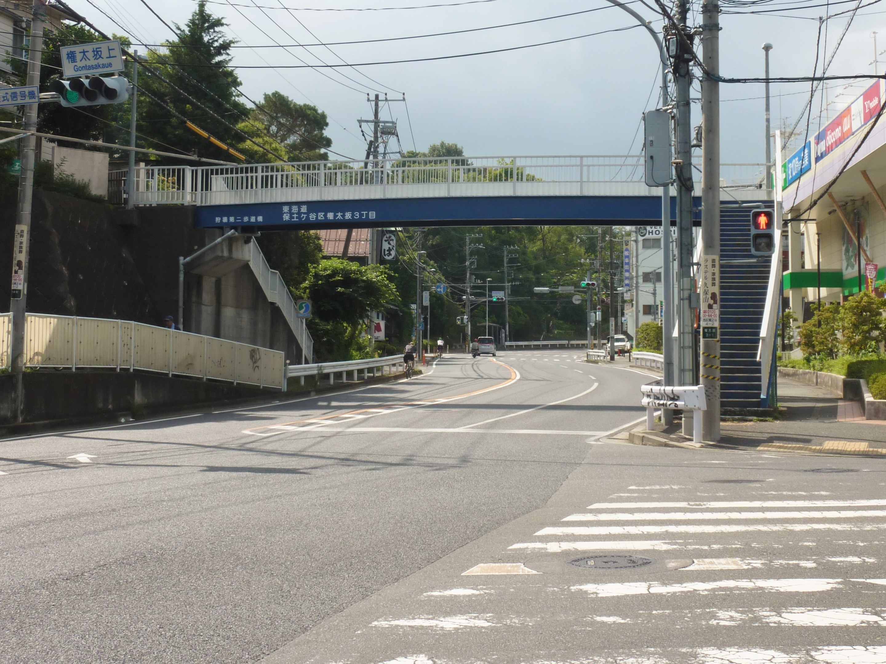 Stadtstraße mit einer Fußgängerbrücke darüber, Fahrzeuge auf der Straße, Strommasten und -leitungen, Verkehrszeichen, Schilder, Gebäude mit Fenstern, Bäume, Pflanzen und Himmel im Hintergrund.