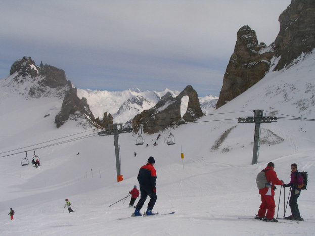 Menschen in Pullovern fahren auf dem Eis Ski, mit einer Seilbahn, Bergen und einem bewölktem Himmel im Hintergrund.