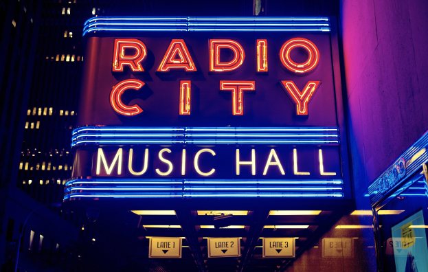 Hell erleuchtetes Radio City Music Hall in New York City mit hohen Gebäuden im Hintergrund und einem "Radio City"-Schild auf der rechten Seite.