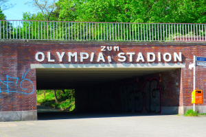 Der Eingang zum Olympiastadion in Berlin, Deutschland, mit einer Brücke mit Text, einem Metallzaun, einem Schild, einer Box, einigen Pflanzen, Gras, einer Gruppe von Bäumen und einem bewölkten Himmel.