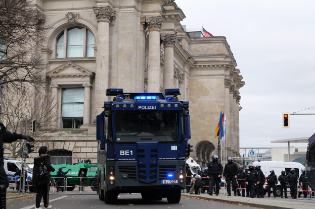 Polizisten vor einem großen Gebäude mit Fahrzeugen auf der Straße, eine Person mit einer Kamera, Bäume, Verkehrszeichen, Flaggen und einem klaren blauen Himmel.