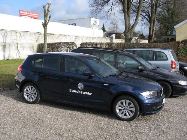 Mehrere Autos sind auf einem von grünem Gras, Büschen und Bäumen umgebenen Weg geparkt, mit einem Schild im Hintergrund, auf dem Text und ein Logo zu sehen sind, sowie Gebäuden.