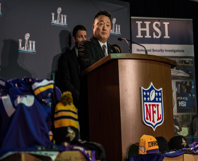 Ein Mann in einem schwarzen Blazer, wei├čem Hemd und schwarzer Krawatte spricht an einem Podium mit einem Mikrofon und einem Logo, gewandt einer Menge; blaue und gelbe Merchandising-Artikel und eine "Super Bowl LII"-Banner sind im Hintergrund sichtbar.