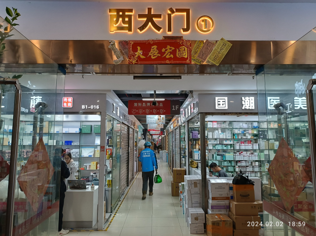 Innenansicht einer chinesischen Apotheke mit Glaswänden, Regalen mit Artikeln, verstreuten Kartons, einer Pflanze links und einer Namensschilder oben.