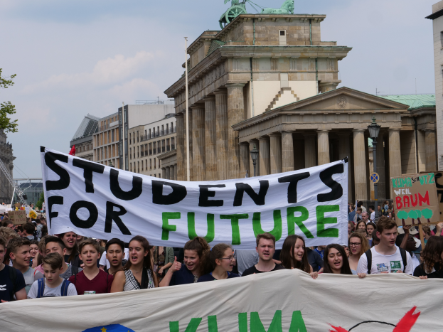 Eine Gruppe von Studenten marschiert in Berlin, wobei sie ein buntes Banner mit der Aufschrift 'Students for Future' tragen, mit Gebäuden, Bäumen und Himmel im Hintergrund.