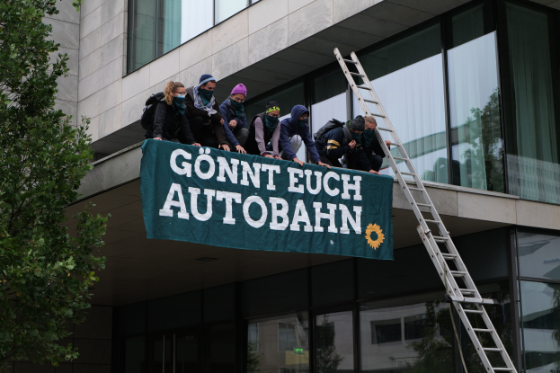 Eine Gruppe von Menschen auf einem Gebäude mit einer Fahne, einem Baum auf der linken Seite und Glasfenstern im Hintergrund, wahrscheinlich protestierend für die deutsche Regierung.