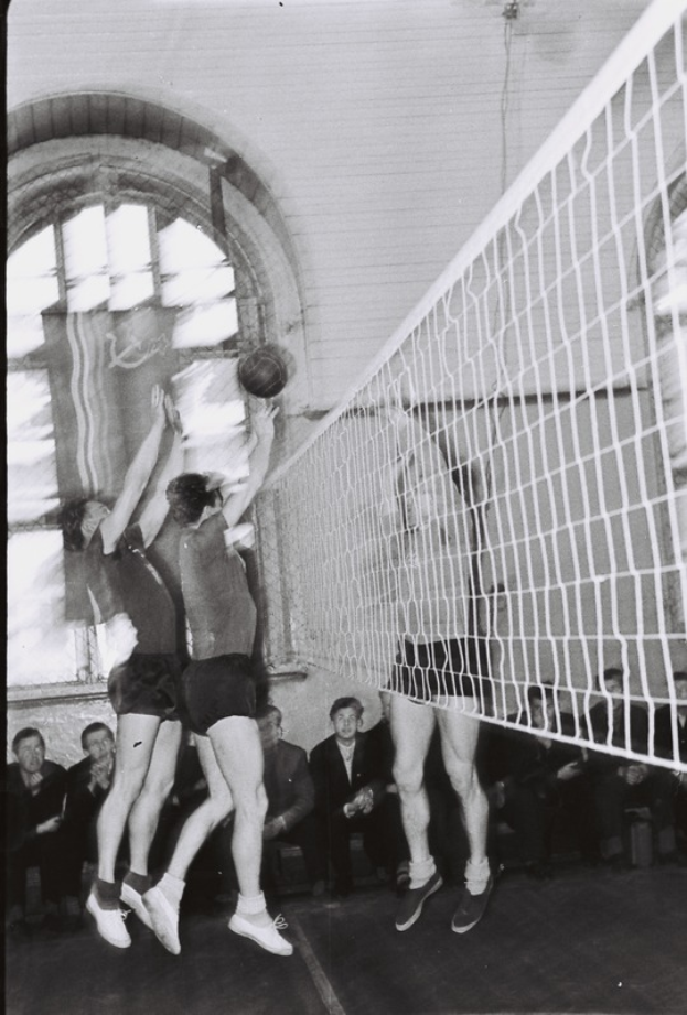 Eine Gruppe von Menschen, die Volleyball in einer Turnhalle mit einem Netz in der Mitte und Zuschauern im Hintergrund spielt, dargestellt in Schwarz-Weiß.