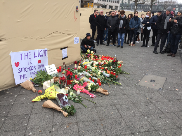 Eine Gruppe von Menschen steht vor einem Zelt mit einer Tafel, auf der "Das Licht ist Stärker als die Dunkelheit" steht, umgeben von Blumensträuschen, Kerzen und anderen Gegenständen, mit Gebäuden, Bäumen und einem klaren Himmel im Hintergrund.