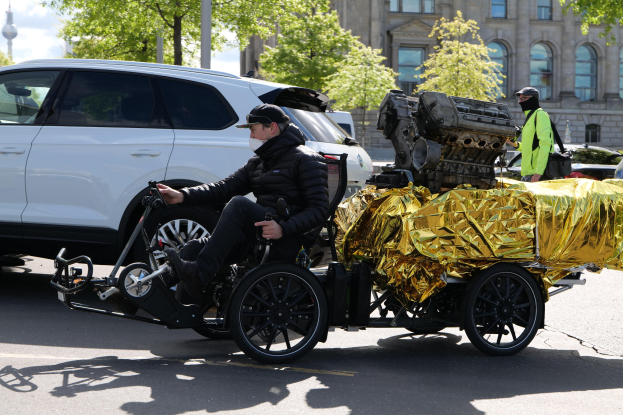 Ein Mann im Rollstuhl mit einem großen Motor auf dem Rücken, umgeben von Fahrzeugen auf einer Straße mit Bäumen, Gebäuden, Polen und einem klaren blauen Himmel im Hintergrund, der eine schwarze Jacke und eine Mütze trägt und ein Objekt hält.
