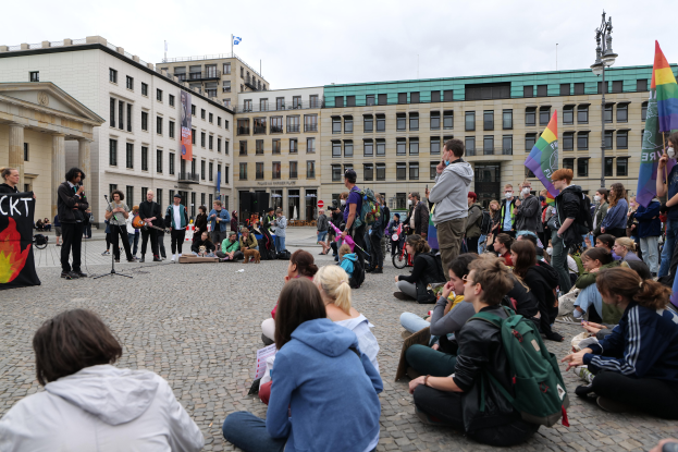 Eine Gruppe von Menschen sitzt auf dem Boden vor einer Menge, die Fahnen und Schilder hält, mit einer Person, die ein Mikrofon hält, einem Schild, einer Statue auf einem Sockel und Gebäuden mit Fenstern im Hintergrund, während einer anti-schwulen Demonstration in Berlin, Deutschland, unter einem bewölkten Himmel.