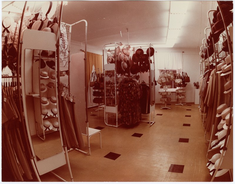 Altes Foto eines Bekleidungsgeschäfts-Innenraums mit Kleiderstangen, Hutständern, einem Spiegel, einem Stuhl, einem Tisch mit Gegenständen, Vorhängen und Deckenleuchten.