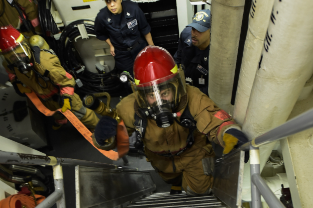 Feuerwehrleute in Schutzausrüstung steigen Treppen mit Rohren und Stangen auf der rechten Seite, verschiedene Gegenstände im Hintergrund.