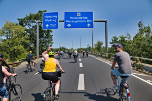 Gruppe von Radfahrern mit Helmen auf einer Straße mit einer Begrenzung auf einer Seite und Bäumen auf der anderen, Laternen und einem klaren blauen Himmel im Hintergrund, mit einem Schild, das eine Radtour in Hamburg anzeigt.