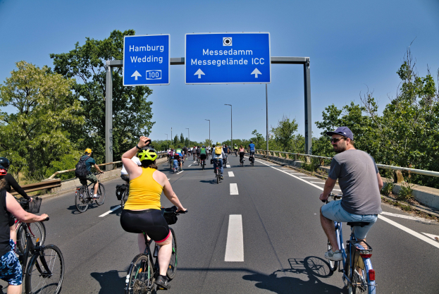 Gruppe von Radfahrern mit Helmen auf einer Straße mit einer Begrenzung auf einer Seite und Bäumen auf der anderen, Laternen und einem klaren blauen Himmel im Hintergrund, mit einem Schild, das eine Radtour in Hamburg anzeigt.