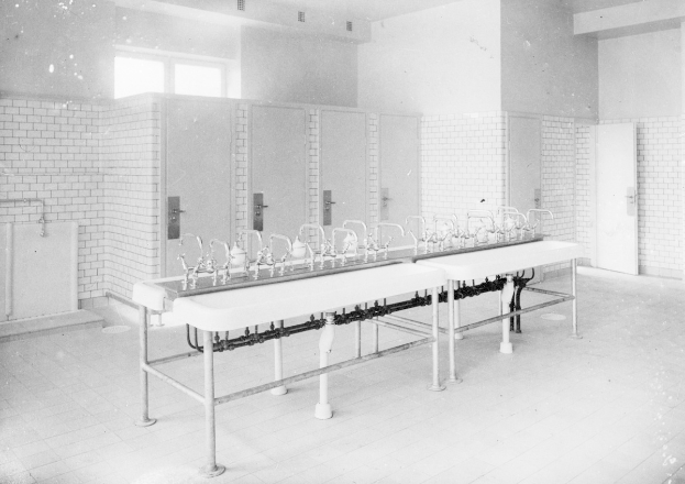 Schwarz-weiß-Foto einer öffentlichen Toilette mit einer Reihe Waschbecken, Wasserhähnen, Wänden, Türen, Fenstern und einem gefliesten Boden, mit verschiedenen Gegenständen, die herumliegen.