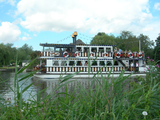 Ein Boot auf einem Teich mit ein paar Menschen darin, umgeben von Gras, Bäumen und einem bewölkten Himmel.