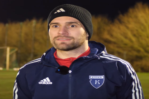 Ein Mann in einer blauen Jacke und Mütze steht auf einem Fußballfeld, umgeben von Bäumen und einem Torpfosten im Hintergrund während eines Trainingssitzung.