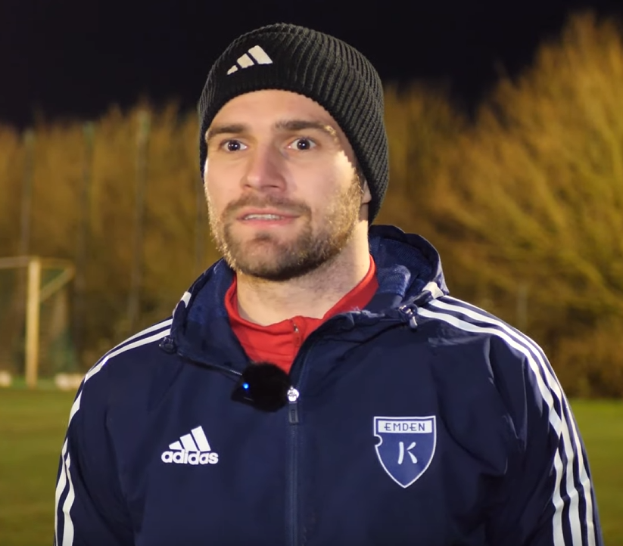 Ein Mann in einer blauen Jacke und Mütze steht auf einem Fußballfeld, umgeben von Bäumen und einem Torpfosten im Hintergrund während eines Trainingssitzung.