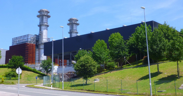 Großes Industriegebäude mit vielen Fenstern, als Kraftwerk identifiziert, umgeben von Straßeninfrastruktur, Fahrzeugen, Vegetation und einem bewölkten Himmel.