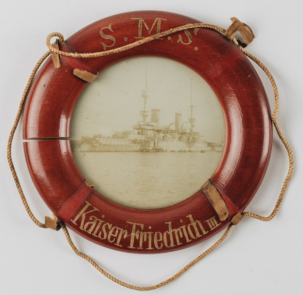 Roter Rettungsring mit 'USS Kaiser Friedrich III'-Text und Bild eines Schlachtschiffs, an einer weißen Oberfl├Ąche mit Seilen befestigt.