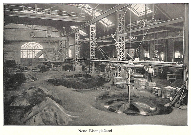 Schwarze und weiße Fotografie einer Fabrik mit Arbeitern, die Maschinen bedienen, umgeben von Metallstangen und -pfählen, beschriftet mit "Neue Eisenglebeerei, die erste Fabrik in Deutschland" unten.