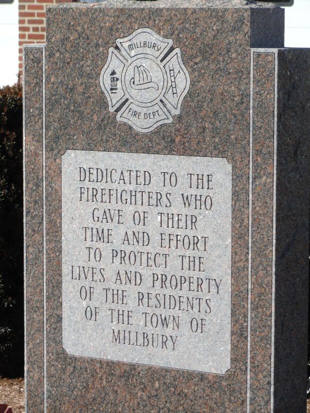Denkmalstein mit einer Gedenktafel für Feuerwehrleute in Millbury vor einem Backsteingebäude mit umgebender Vegetation.