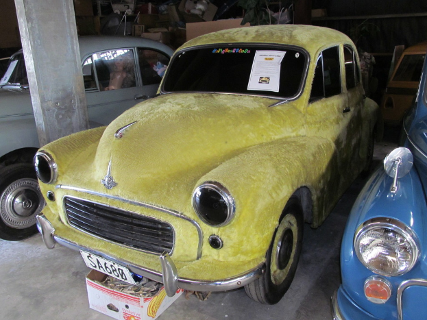 Mehrere Oldtimer sind vor einer Wand mit verschiedenen Zubehörteilen geparkt, wobei auf der linken Seite eine Säule zu sehen ist.