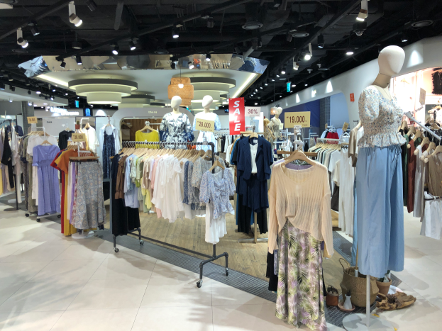 Ein Bekleidungsgeschäft mit Schaufensterpuppen in Kleidern, Kleidung an Bügeln, Schautafeln mit Text, Deckenbeleuchtung und verschiedenen Gegenständen auf dem Boden, was darauf hinweist, dass es ein Modegeschäft ist.