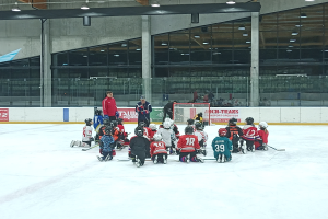 Eine Gruppe von Kindern in Helmen und mit Hockey-Schlägern sitzt auf einem Eislaufplatz, mit einer Wand aus Glas und Säulen, Deckenleuchten und Texttafeln im Hintergrund.