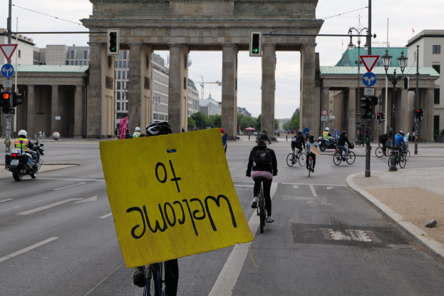 Eine Gruppe von Radfahrern mit Helmen fährt auf einer Straße am Brandenburger Tor in Berlin, Deutschland, vorbei, wobei einer ein gelbes Schild hält, vor einem klaren blauen Himmel mit Gebäuden und Bäumen im Hintergrund.
