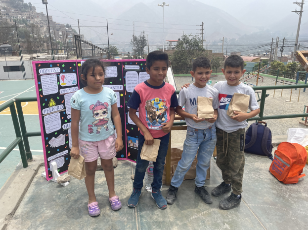 Gruppe von Kindern auf einem Basketballfeld, die Papierbeutel halten, mit Beuteln und Gegenständen rechts daneben und einer Tafel mit Text links daneben, vor einem Hintergrund aus Zaun, Pfosten, Straßenlaternen, Strommasten, Bäumen, Gebäuden, Hügeln und Himmel.
