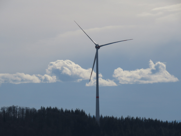 Windkraftanlage in einer bewaldeten Gegend mit Hügeln und bewölktem Himmel im Hintergrund.