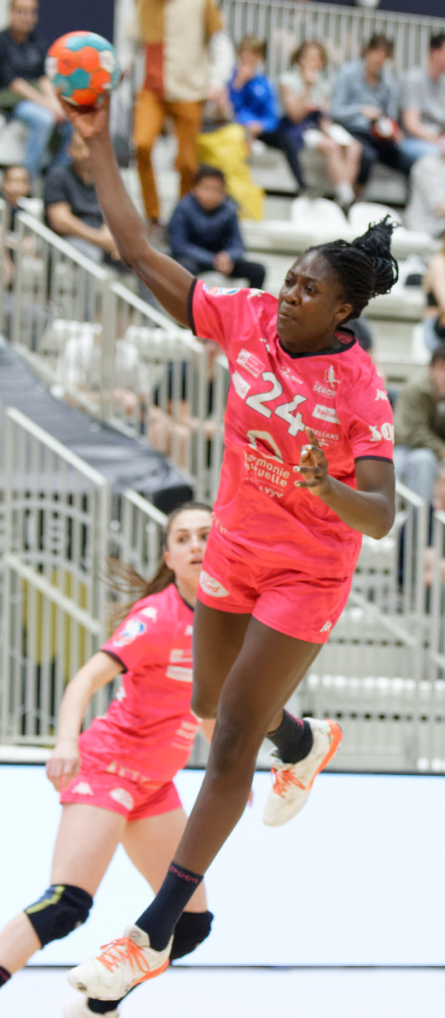 Zwei Frauen in Sportuniformen spielen Handball vor einem sitzenden und stehenden Publikum mit Geländern im Hintergrund.
