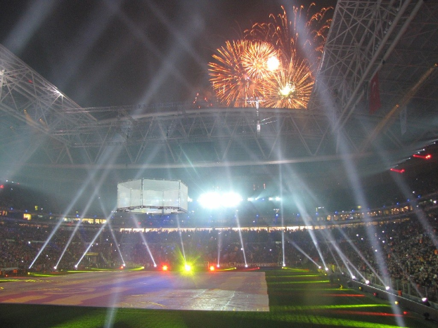 Olympiastadion in London, England, bei Nacht mit grüner Wiese, umgeben von einer Menge und Feuerwerk am Himmel.