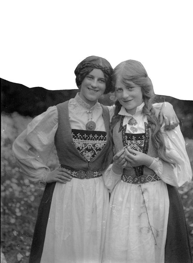 Zwei junge Frauen in traditioneller bayrischer Tracht, mit weißen und schwarzen Kleidern mit aufwendiger Stickerei und Perlen, posieren vor einer Kulisse aus üppigen grünen Pflanzen und einem hellen Himmel.