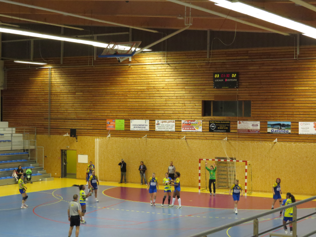 Eine Gruppe von Menschen, die in einer Turnhalle Handball spielen, mit einem Metallzaun, Torpfosten, Tür, Texttafeln, Anzeigebildschirm, Lautsprecherbox, Metallstangen und einem Dach mit Deckenleuchten und Metallrahmen.
