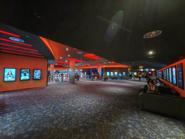 Großer Raum mit Menschen auf Sofas sitzend, die Wandfernseher beobachten, umgeben von Säulen und Deckenleuchten, mit sichtbarem Boden.