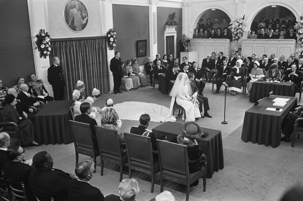 Ein Schwarz-Weiß-Foto einer Hochzeitszeremonie in einem großen Raum mit Gästen auf Stühlen und Stehplätzen, Tischen mit Papieren und Mikrofonen, einer dekorierten Wand mit Fotorahmen und Blumenbouquets sowie einem Podium mit Mikrofonständer.