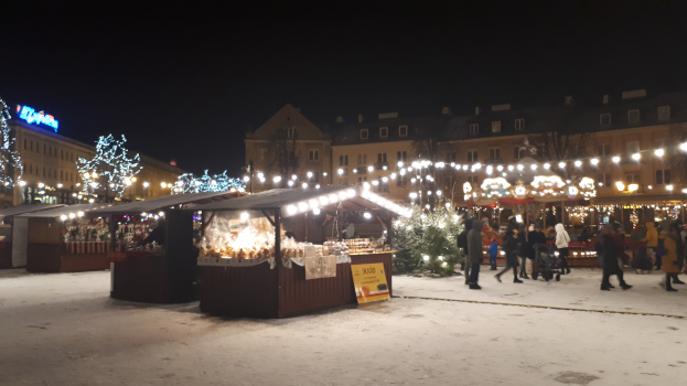 Ein Weihnachtsmarkt in einer verschneiten Straße bei Nacht, mit Menschen, Kinderwagen, beleuchteten Hütten, Pflanzen, Bäumen, Gebäuden und Schildern.